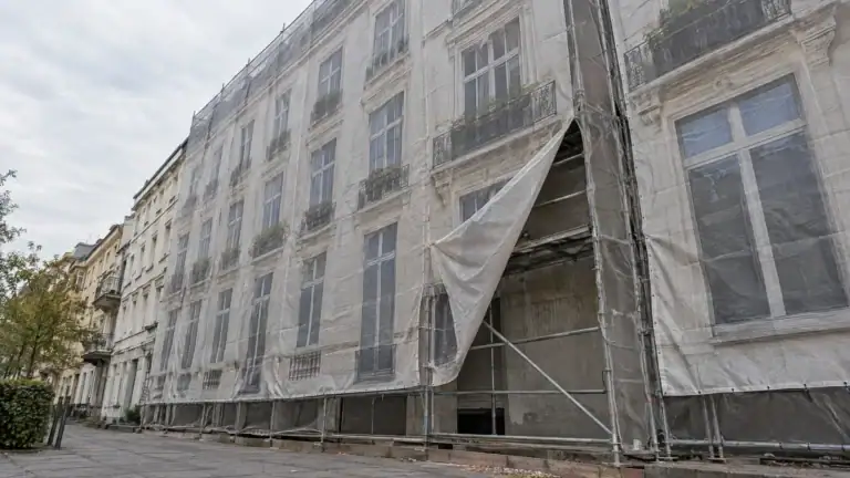 Mit Gerüstplane verhüllte Altbaufassade, an einer Stelle aufgerissen, dahinter Rohbau und Gerüst (Symbolbild, KI-generiert)