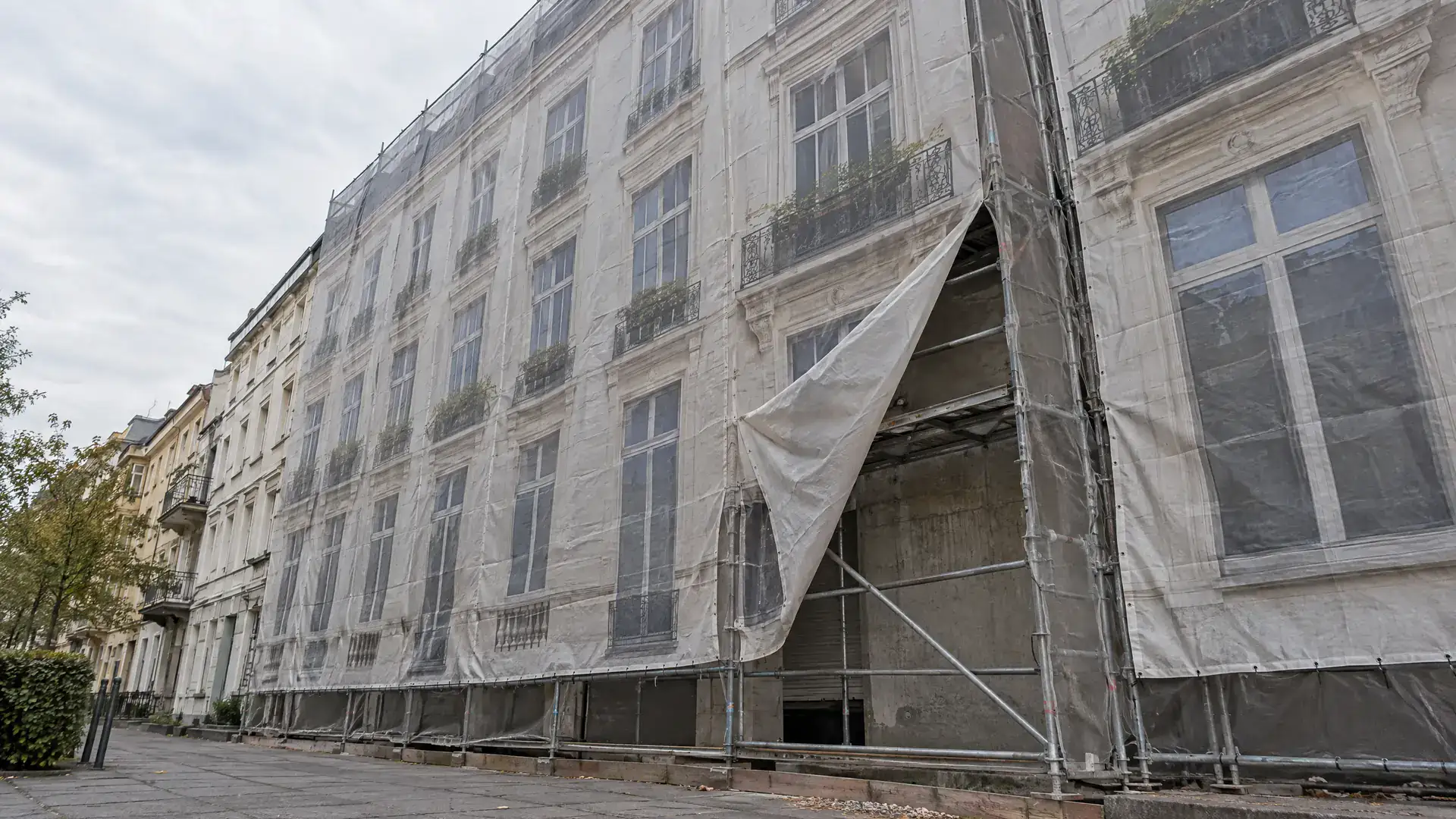 Mit Gerüstplane verhüllte Altbaufassade, an einer Stelle aufgerissen, dahinter Rohbau und Gerüst (Symbolbild, KI-generiert)