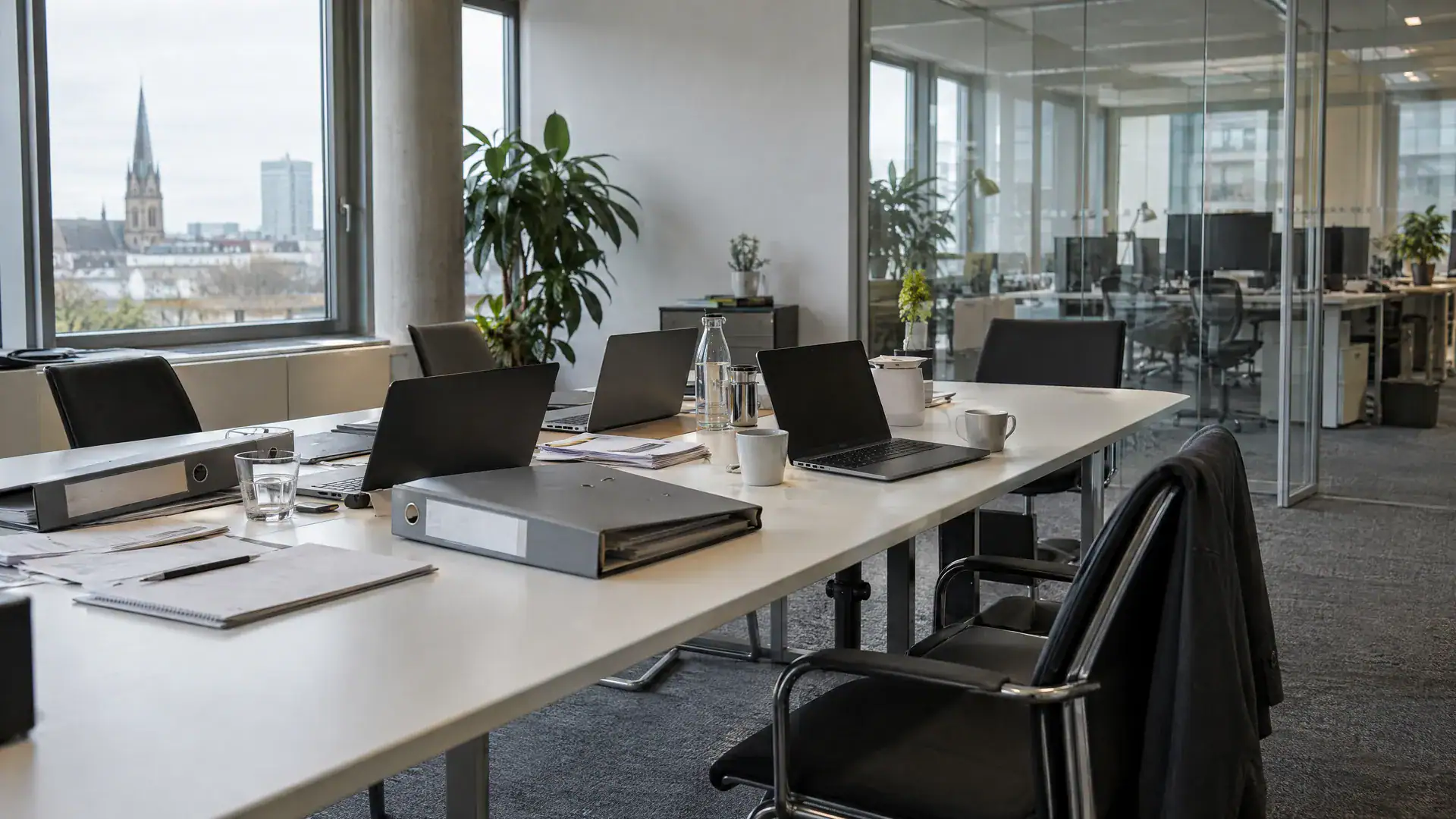 Prüferraum beim Mandanten mit langem Tisch, aufgeklappten Laptops, Kaffeetassen und Glaswand (Symbolbild, KI-generiert)