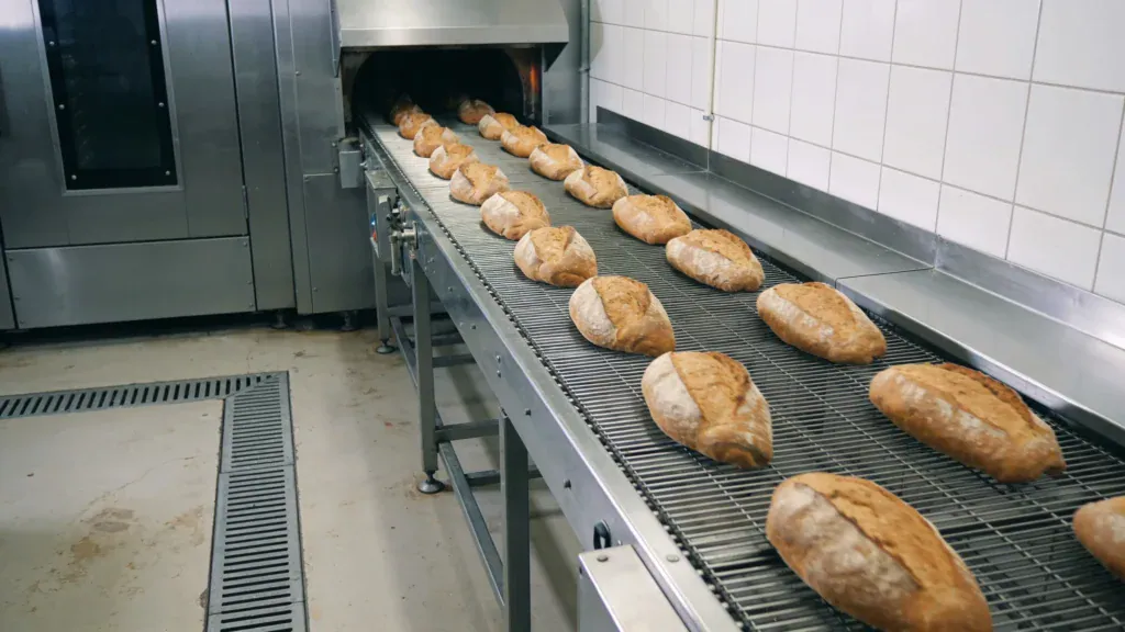 Großbäckerei, Brote laufen aus dem Tunnelofen auf ein Gitterförderband (Symbolbild, KI-generiert)