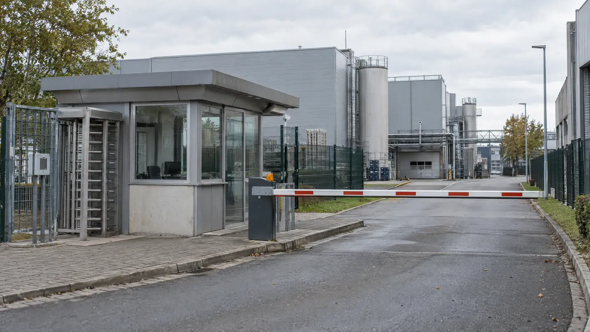 Werkszufahrt mit Pförtnerhaus, Schranke und Werkshallen im Hintergrund (Symbolbild, KI-generiert)