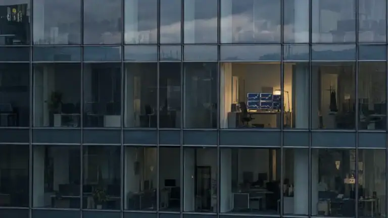 Dunkle Glasfassade eines Bürohochhauses, ein erleuchteter Raum mit mehreren Bildschirmen (Symbolbild, KI-generiert)