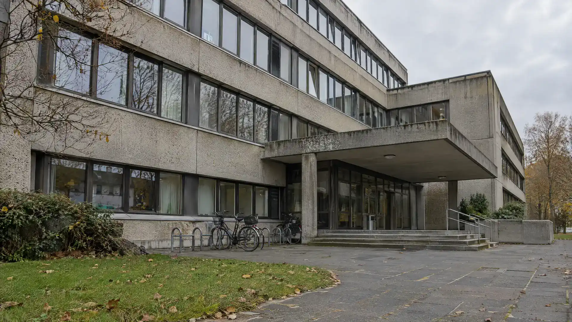 Behördengebäude aus den Siebzigern mit Betonfassade, Fahrradständern und Eingangsvordach (Symbolbild, KI-generiert)