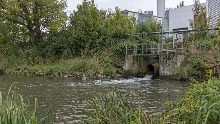 Einleitungsrohr eines Werks an einem kleinen Fluss mit Probenahmesteg (Symbolbild, KI-generiert)