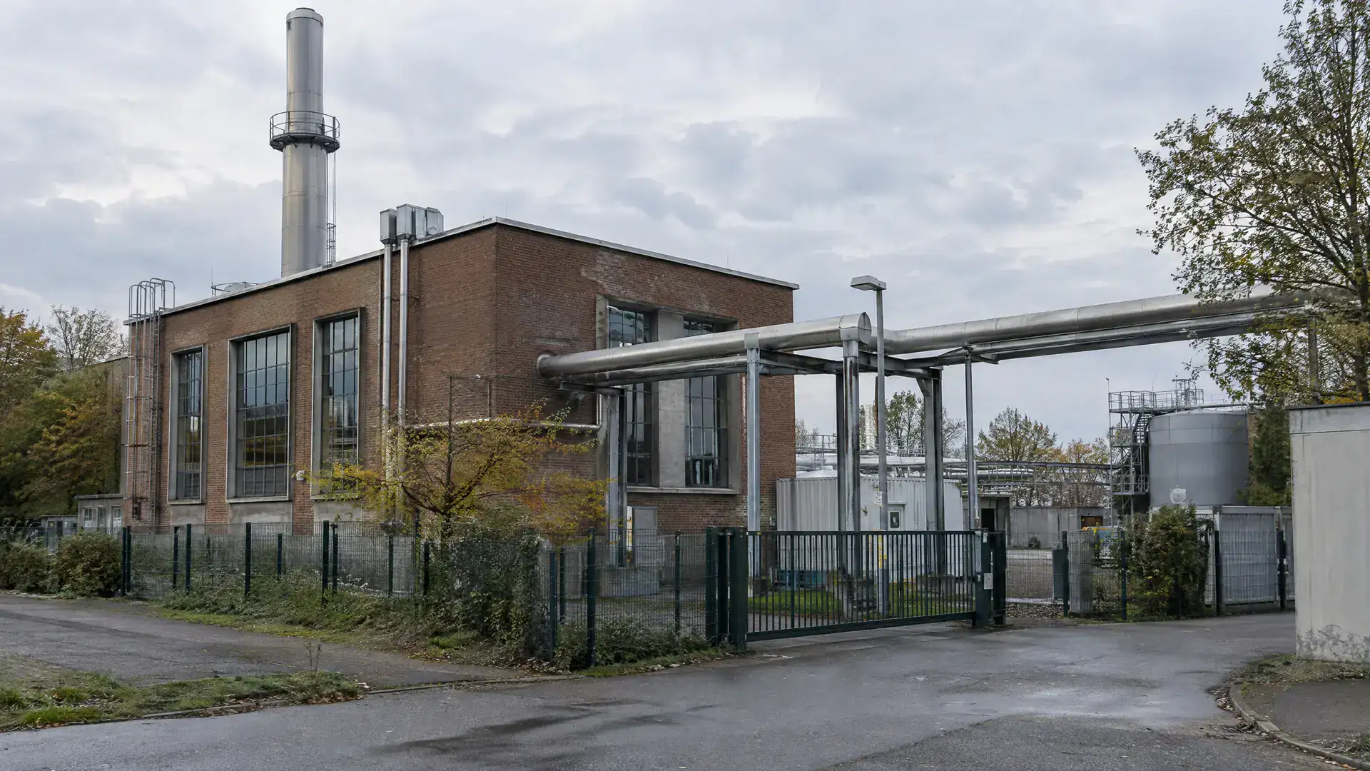 Kleines städtisches Heizkraftwerk mit Backsteinbau, Schornstein und Fernwärmeleitungen (Symbolbild, KI-generiert)